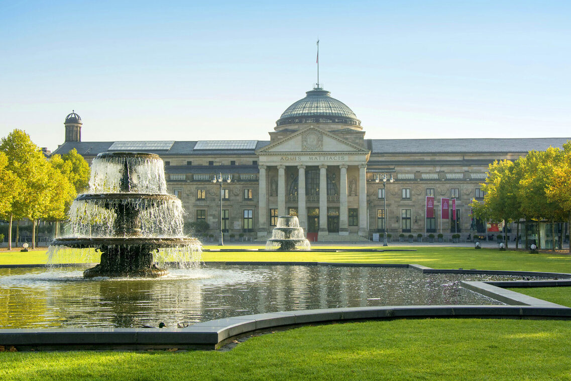 Historisches Gebäude mit Springbrunnen und viel Grün.